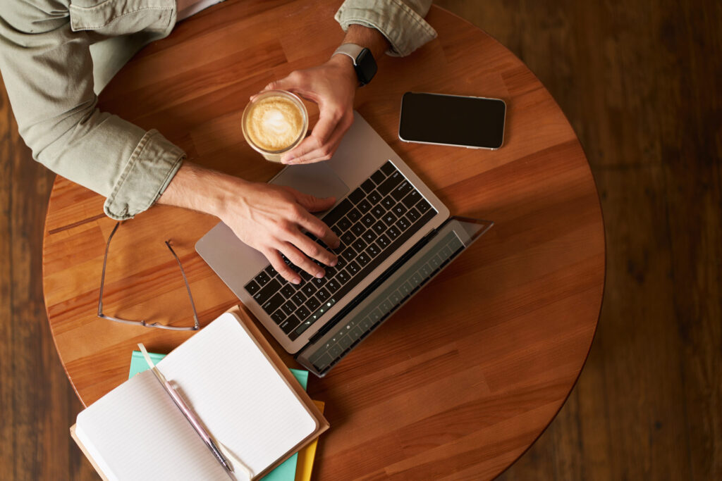 co working space, freelance and e learning concept. top view, male hands holding cup of coffee and using laptop. a man sitting in cafe and working or studying online